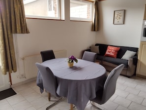 A dining area with a table, chairs, a vase with flowers, a radiator, a sofa, and a window with curtains. - Gîte rural Le Vent du Nord – Ferme des 4 Vents (Audinghen)