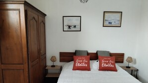 A bedroom with a large wooden wardrobe, a bed with white bedding, two red pillows with French text, a small bedside table with a lamp, and two framed pictures on the wall. - Gîte rural Le Vent du Nord – Ferme des 4 Vents (Audinghen)