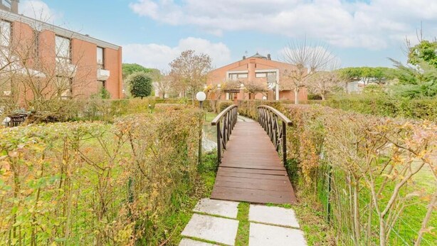 Eine Holzbrücke führt zu einem Haus mit Ziegelverkleidung, umgeben von viel Grün und blauem Himmel.