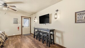 A room with a wooden floor, a ceiling fan, a TV mounted on the wall, a bar area with stools, and a door with a window. - Old West Studio Cabin Near Yellowstone (Island Park)