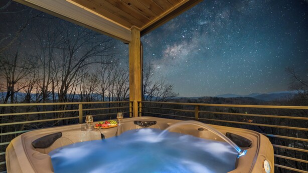 A hot tub with a night sky view, a table with a glass of wine, and a bowl of fruit. - Hot Tub w/ Panoramic Views + Indoor Pool (Sevierville)