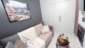 Ein modernes Wohnzimmer mit grauer Wand, einem großen gerahmten Bild, einer Couch mit gemusterten Kissen, einem kleinen Holztisch mit Obst und einem weißen Kleiderschrank.
