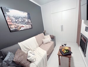 Ein modernes Wohnzimmer mit grauer Wand, einem großen gerahmten Bild, einer Couch mit gemusterten Kissen, einem kleinen Holztisch mit Obst und einem weißen Kleiderschrank.
