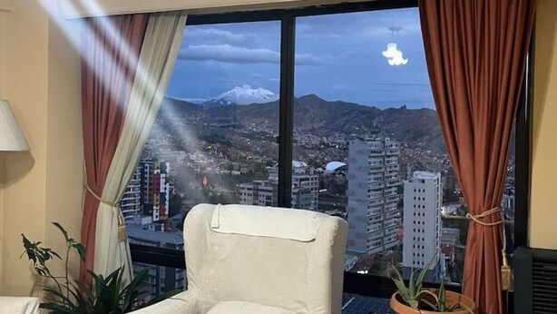 Habitación de hotel con un amplio ventanal que ofrece vistas a un paisaje urbano y montañas.