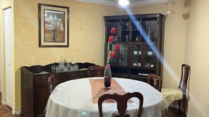 Un comedor con una mesa redonda cubierta por un mantel blanco, rodeada de sillas de madera. Un jarrón con flores rojas está sobre la mesa.