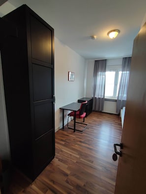 A room with a wooden floor, a desk with a chair, a wardrobe, and a window with curtains.
