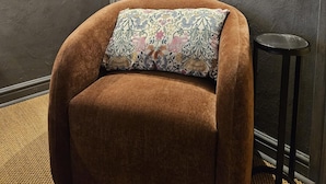 A room with a brown upholstered armchair, a patterned pillow, a framed floral artwork, a door, and a small black stool.