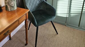 A room with a wooden desk, a patterned lamp, a dark upholstered chair, a woven carpet, and a green door with a geometric design.