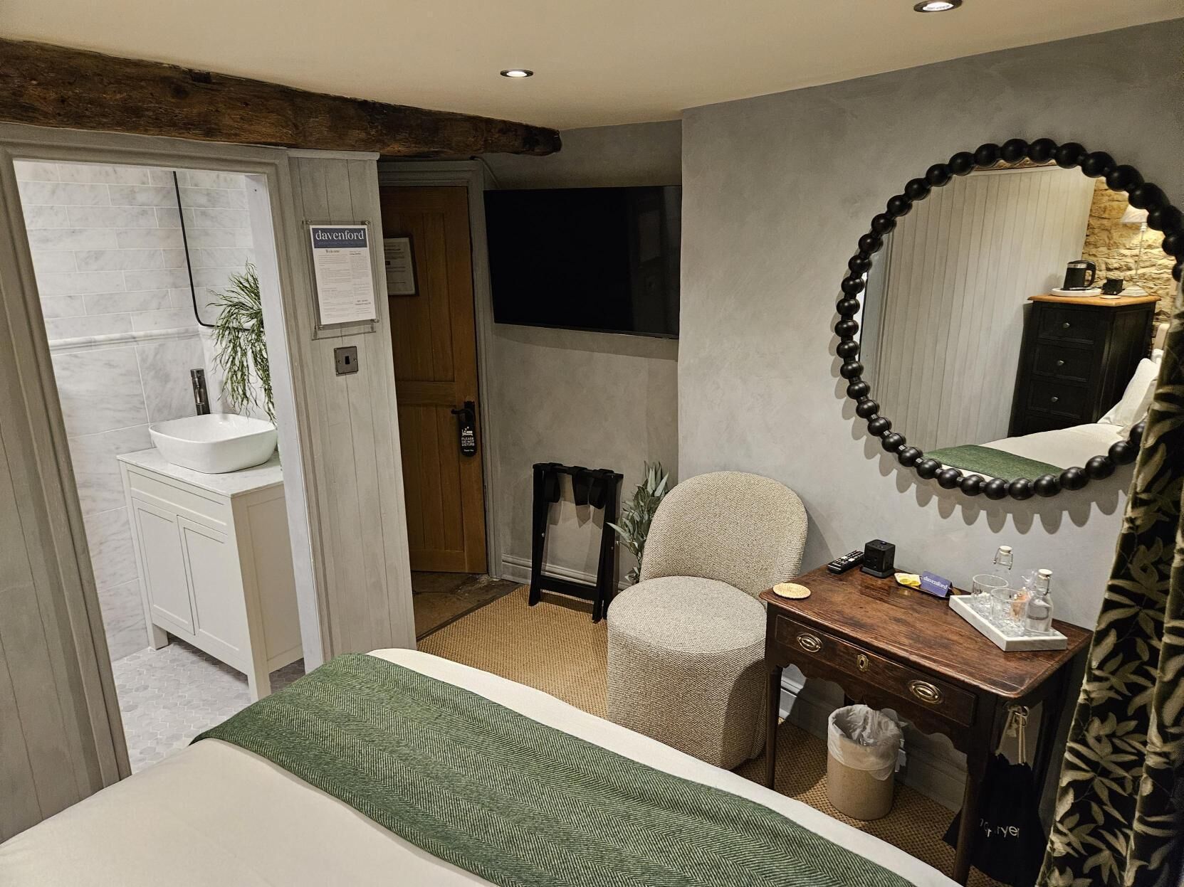 A bedroom with a bed, a bedside table, a chair, a mirror, a dresser, and a bathroom visible through an open door.