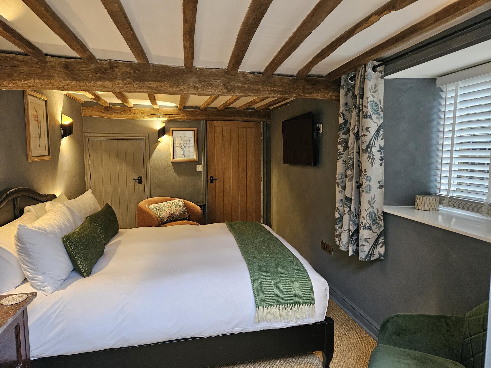 Ein Schlafzimmer mit einem Bett, einem Sessel, einem Fenster mit Jalousien und einer Holzbalkendecke.