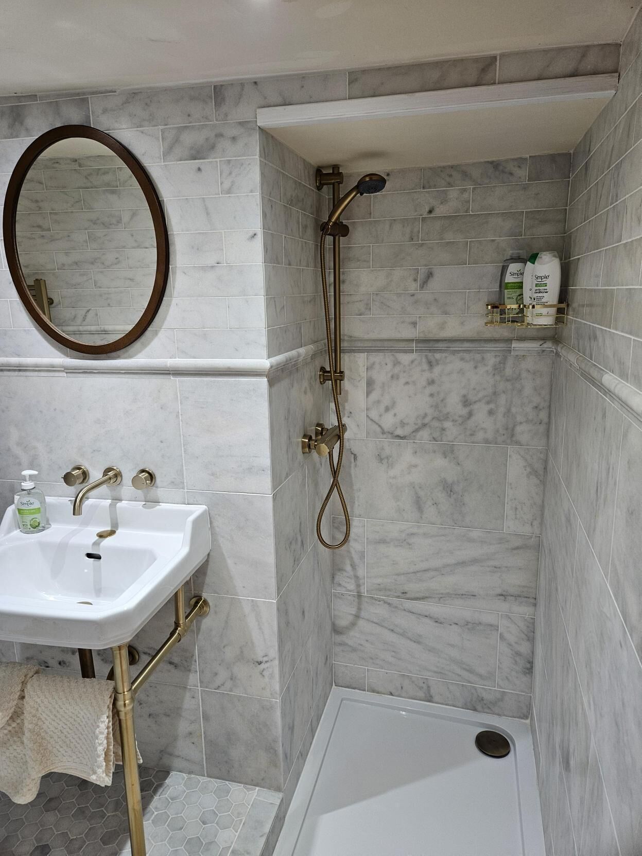 A bathroom with a white sink, a mirror, a shower with a handheld showerhead, and a shelf with toiletries.