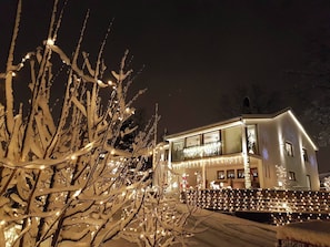 A modern house with a balcony, illuminated by string lights, surrounded by snow-covered trees.