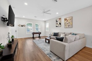 A modern living room with a sofa, coffee table, and a foosball table. There is a flat-screen TV mounted on the wall and a ceiling fan. - Stylish Tampa Home: Complimentary Coffee&Wine Bar (Tampa)