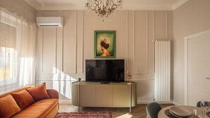 Una sala de estar con un sofá, un televisor sobre un mueble, una araña y un cuadro en la pared.