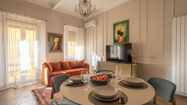 Un comedor con una mesa redonda preparada para cuatro personas, rodeada de sillas. Hay un sofá y un televisor al fondo.