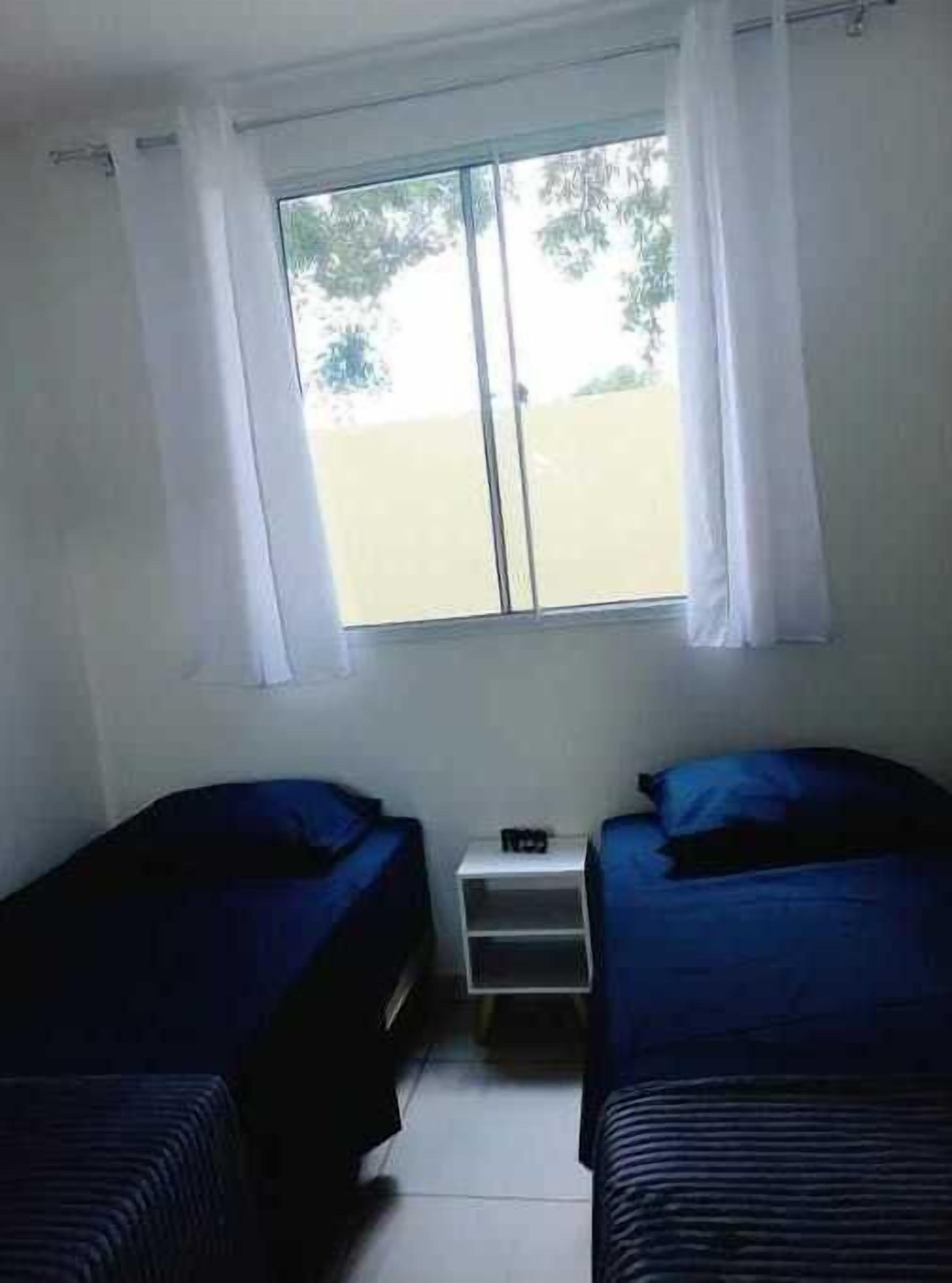 Ein kleines, schlichtes Zimmer mit zwei Einzelbetten, einem kleinen Tisch und einem Fenster mit Vorhängen.