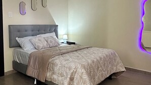 Un dormitorio con cama, mesita de noche, espejo en la pared y lámpara en el techo.