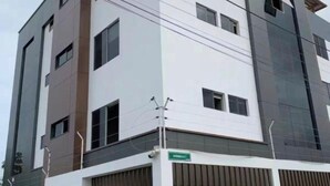 Un edificio de varios pisos con un diseño arquitectónico moderno, que combina paneles blancos y grises oscuros, ventanas grandes y una entrada protegida por una reja.