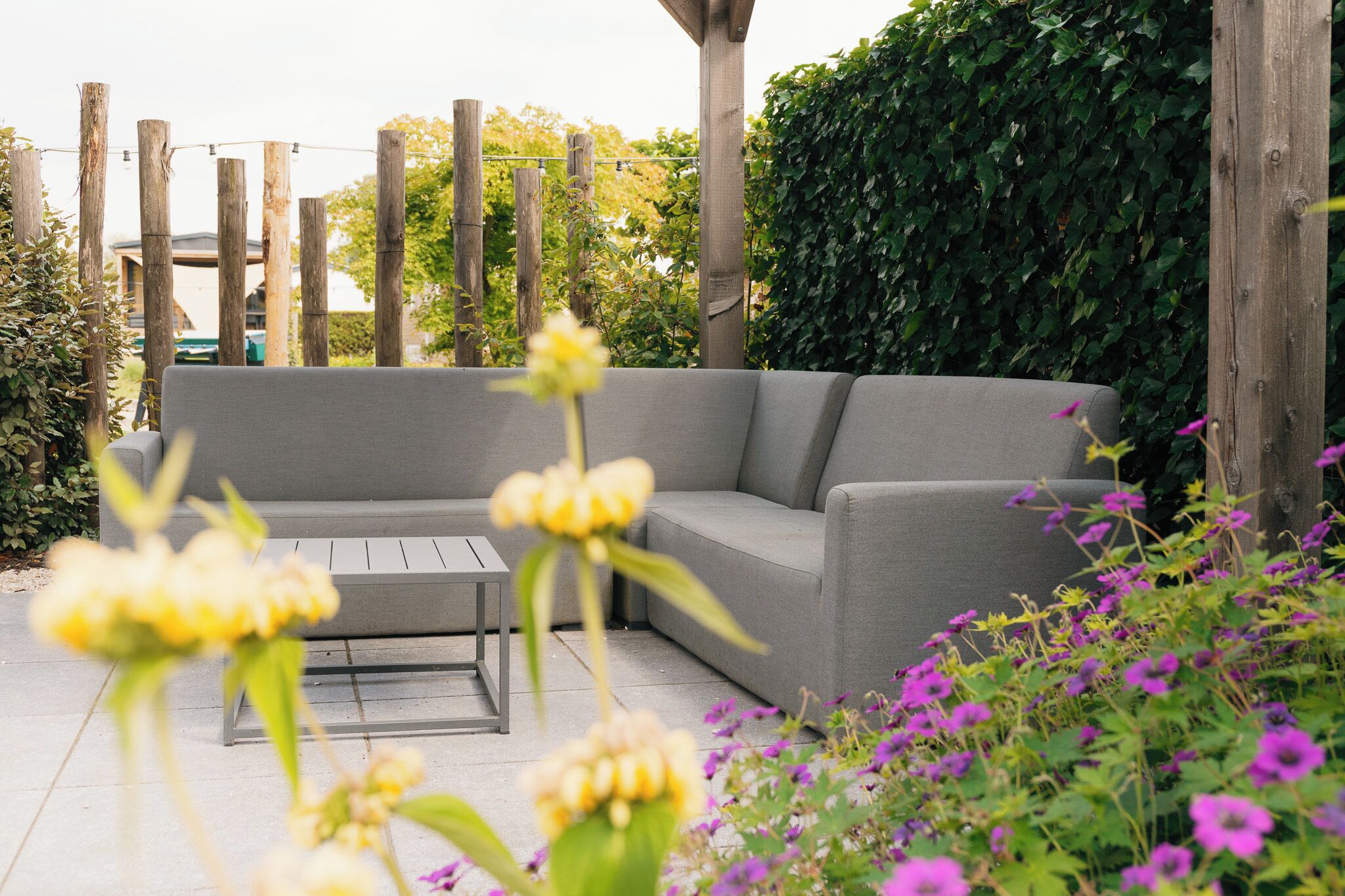 A grey outdoor seating area with a wooden pergola, surrounded by greenery and flowers.