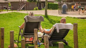 Zwei Personen entspannen auf Liegestühlen in einer parkähnlichen Umgebung mit einem Spielplatz und Gebäuden im Hintergrund.
