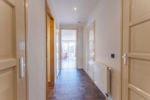 Un couloir avec une porte en bois sur la gauche, une porte blanche sur la droite, et une vue sur une pièce où l’on aperçoit un lit et une fenêtre.