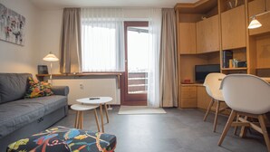 A compact living space with a sofa, a small round table, a colorful ottoman, a kitchenette area, and a dining set. - Appartement - Heidestrasse 2-17 | Winterberg 'Hermanna' (Winterberg)