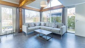 Een moderne woonkamer met een grijze modulaire bank, een glazen salontafel en grote ramen met uitzicht op het landschap.