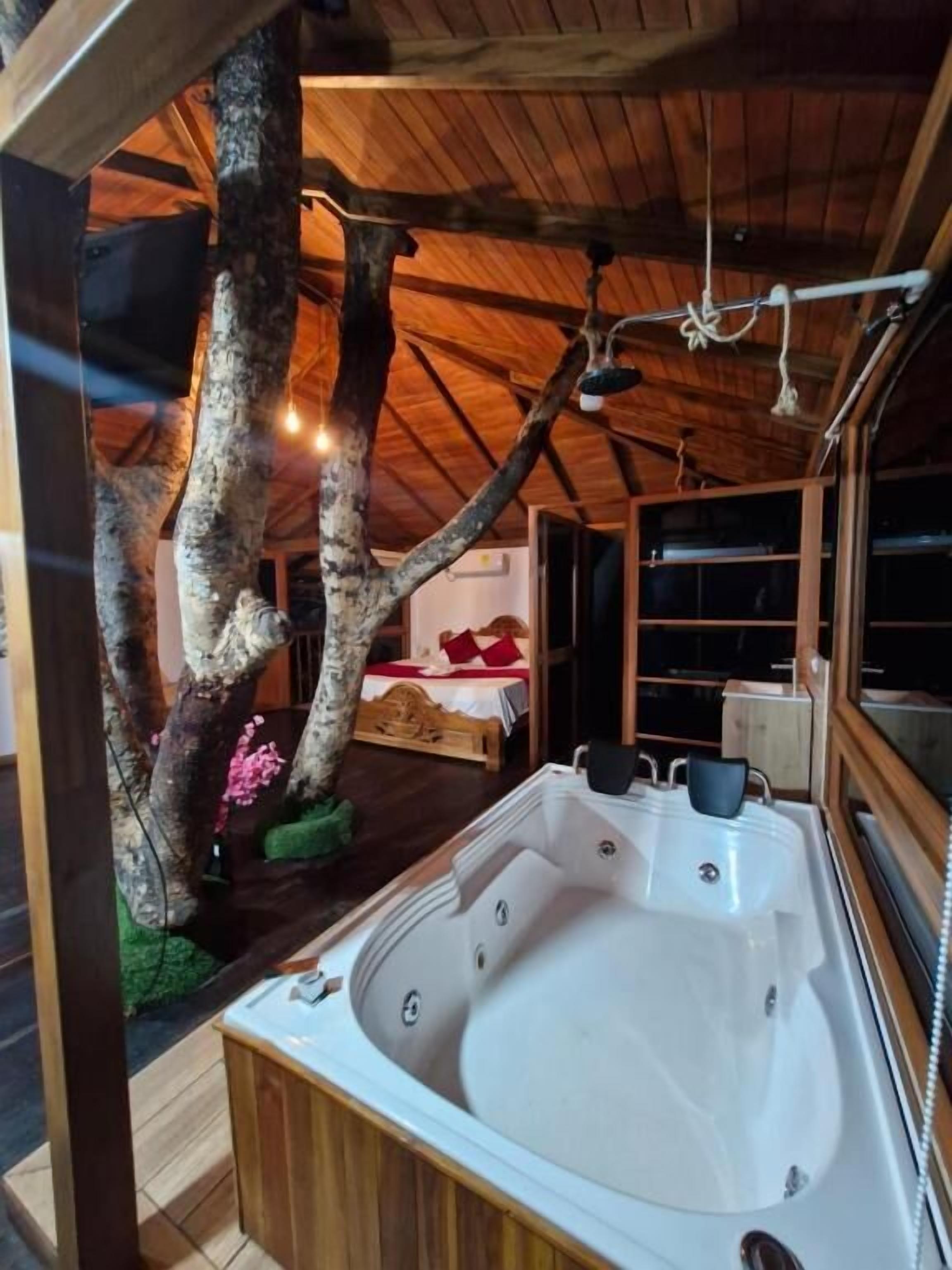 A wooden cabin with a large hot tub, a bed with red bedding, and a tree with pink flowers.
