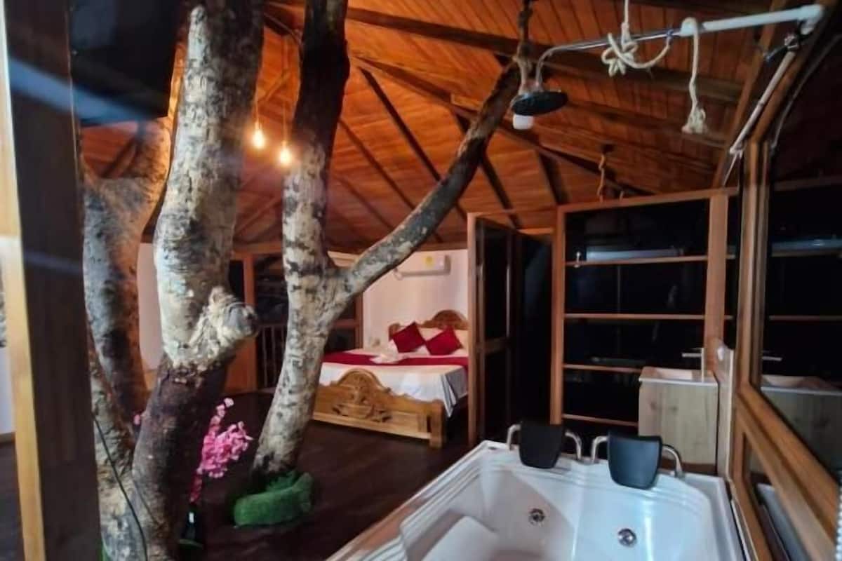 A wooden cabin with a large hot tub, a bed with red bedding, and a tree with pink flowers.