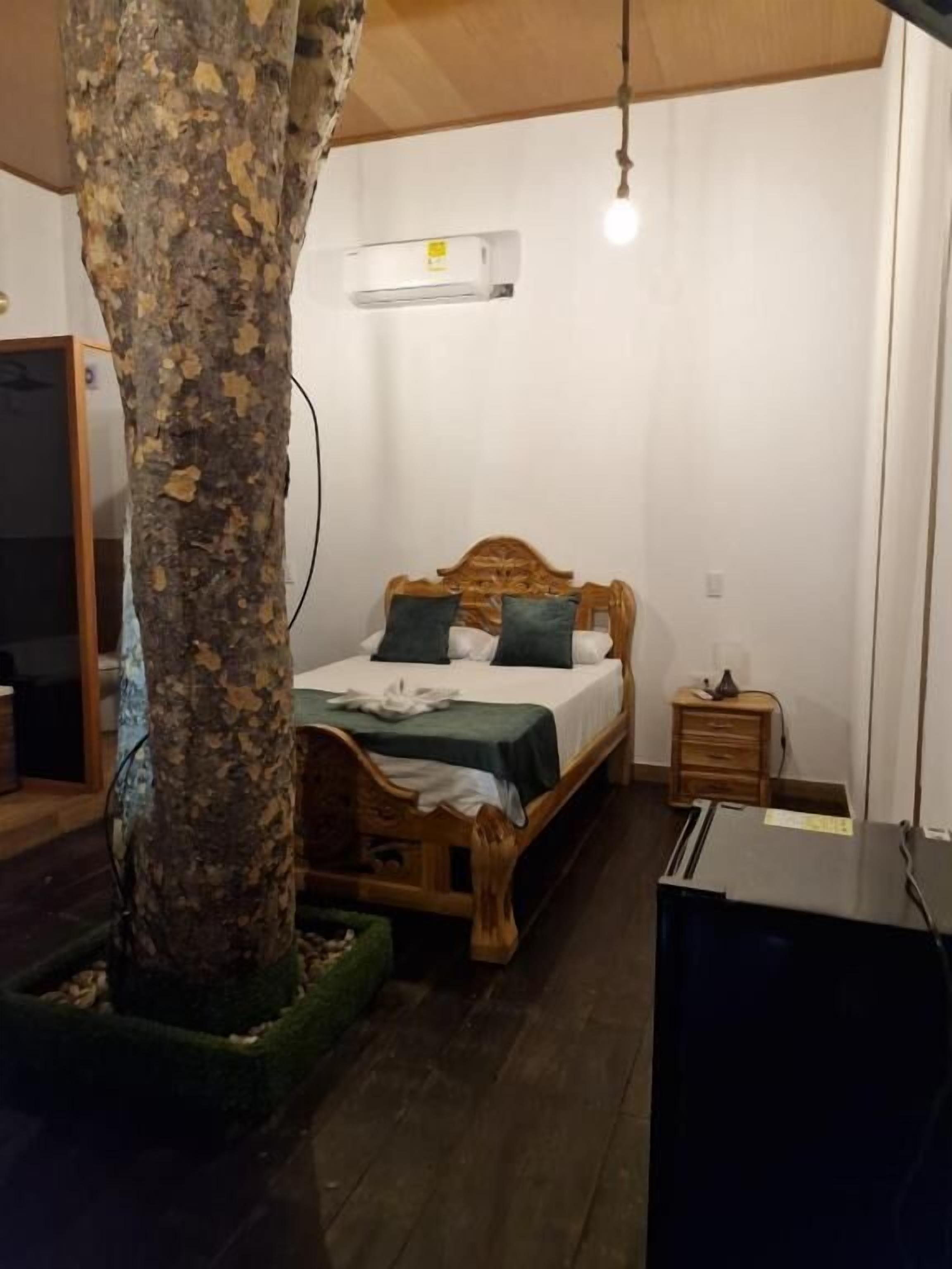 Un dormitorio con una cama de madera, un tronco de árbol, una mesita de noche pequeña y un equipo de aire acondicionado.