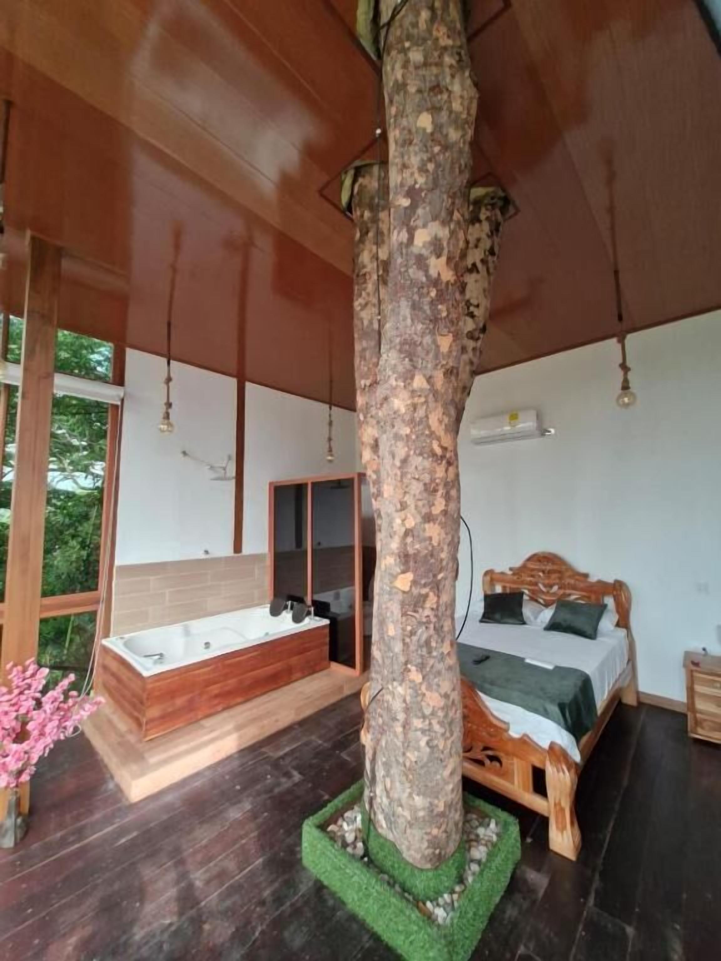 Camera da letto con un grande tronco d'albero al centro, un letto e una panca in legno, una vasca da bagno e un'ampia finestra.