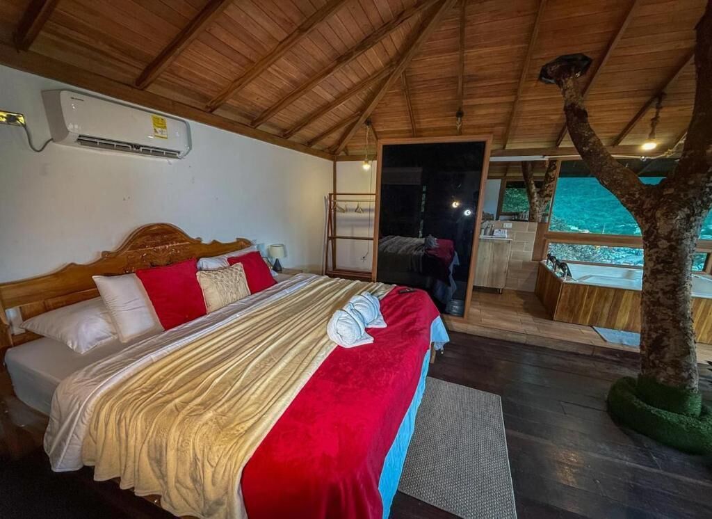 A bedroom with a wooden ceiling, a bed with red and beige bedding, a window, and a tree outside.