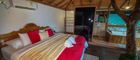 A bedroom with a wooden ceiling, a bed with red and beige bedding, a window, and a tree outside.