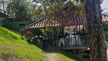 A covered outdoor seating area with tables and chairs, surrounded by greenery and trees.