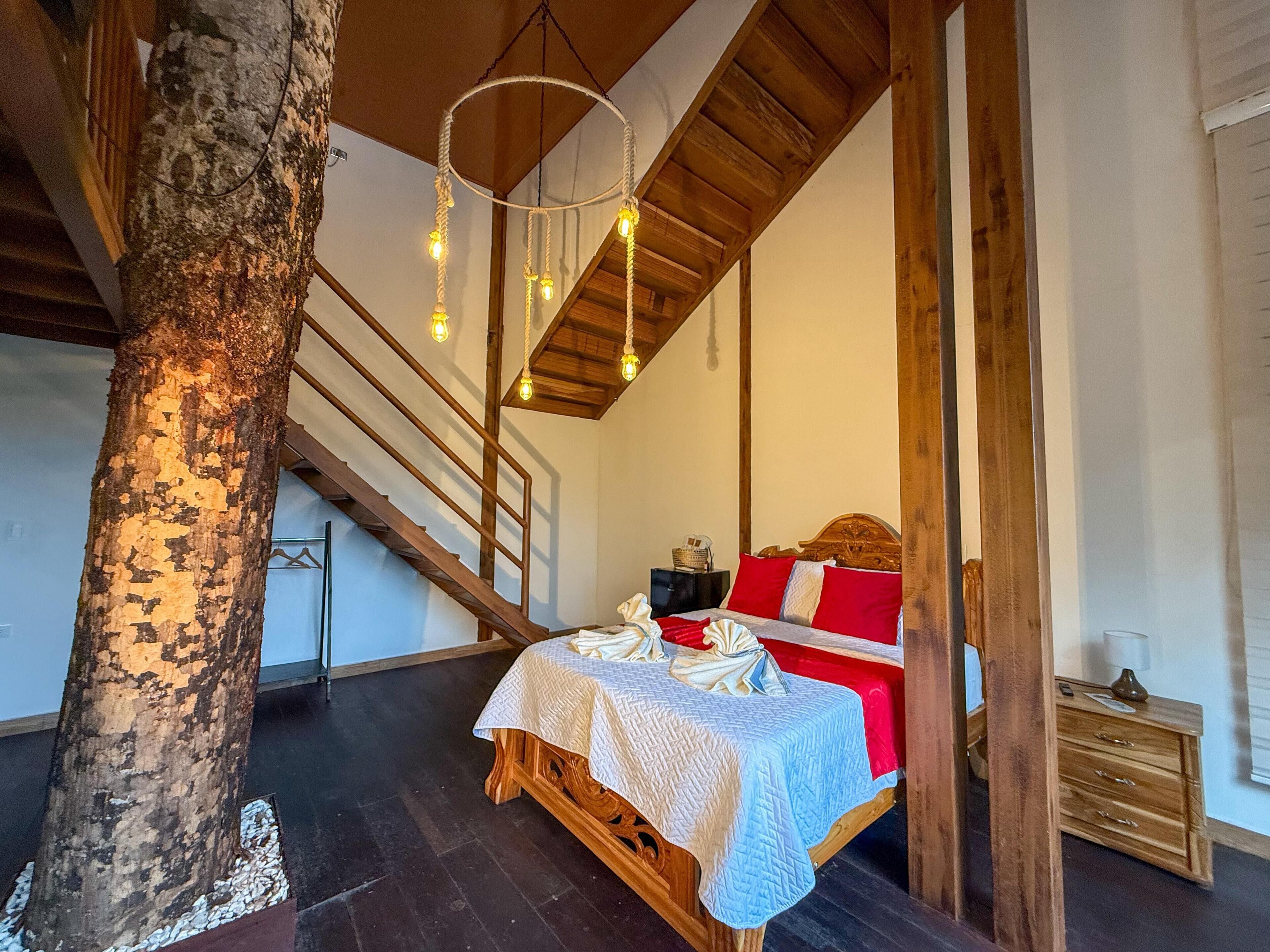 Een slaapkamer met een houten bed, nachtkastje, dressoir, een trap en een boomstam.