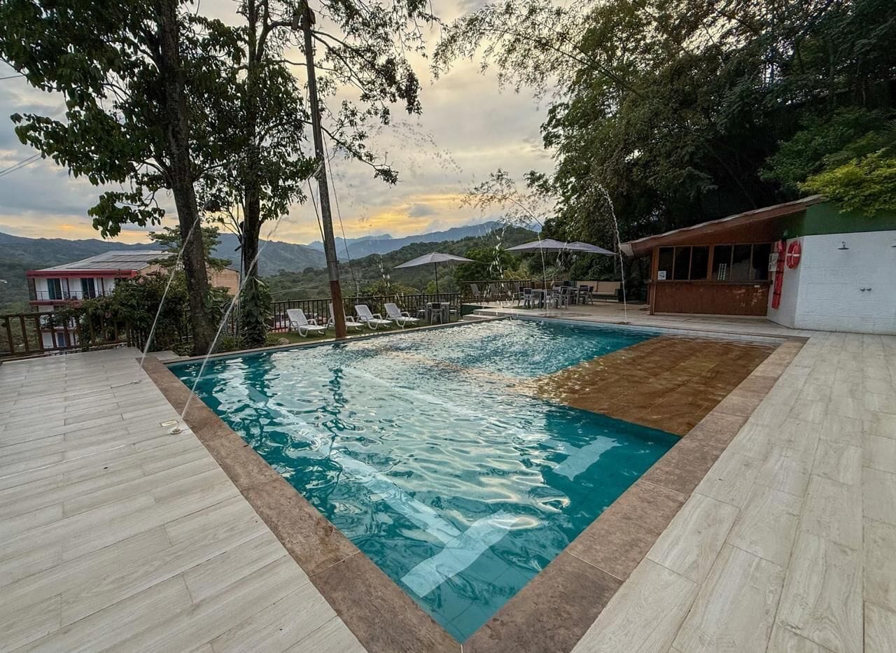 A swimming pool with a wooden deck, surrounded by lounge chairs and umbrellas, with a view of mountains in the background.
