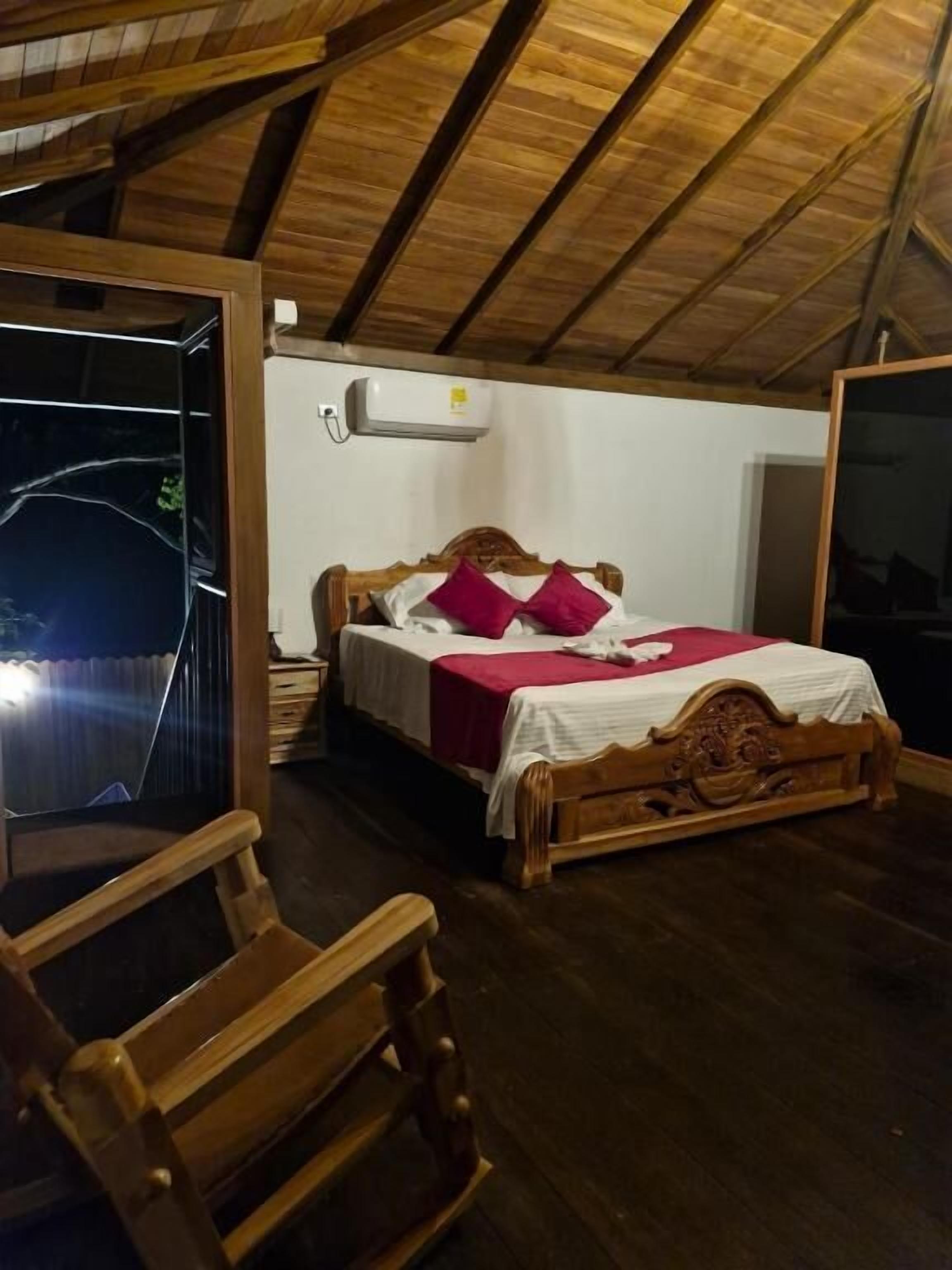Un dormitorio con techo de madera, una cama con colcha blanca y roja, una silla de madera y un equipo de aire acondicionado en la pared.
