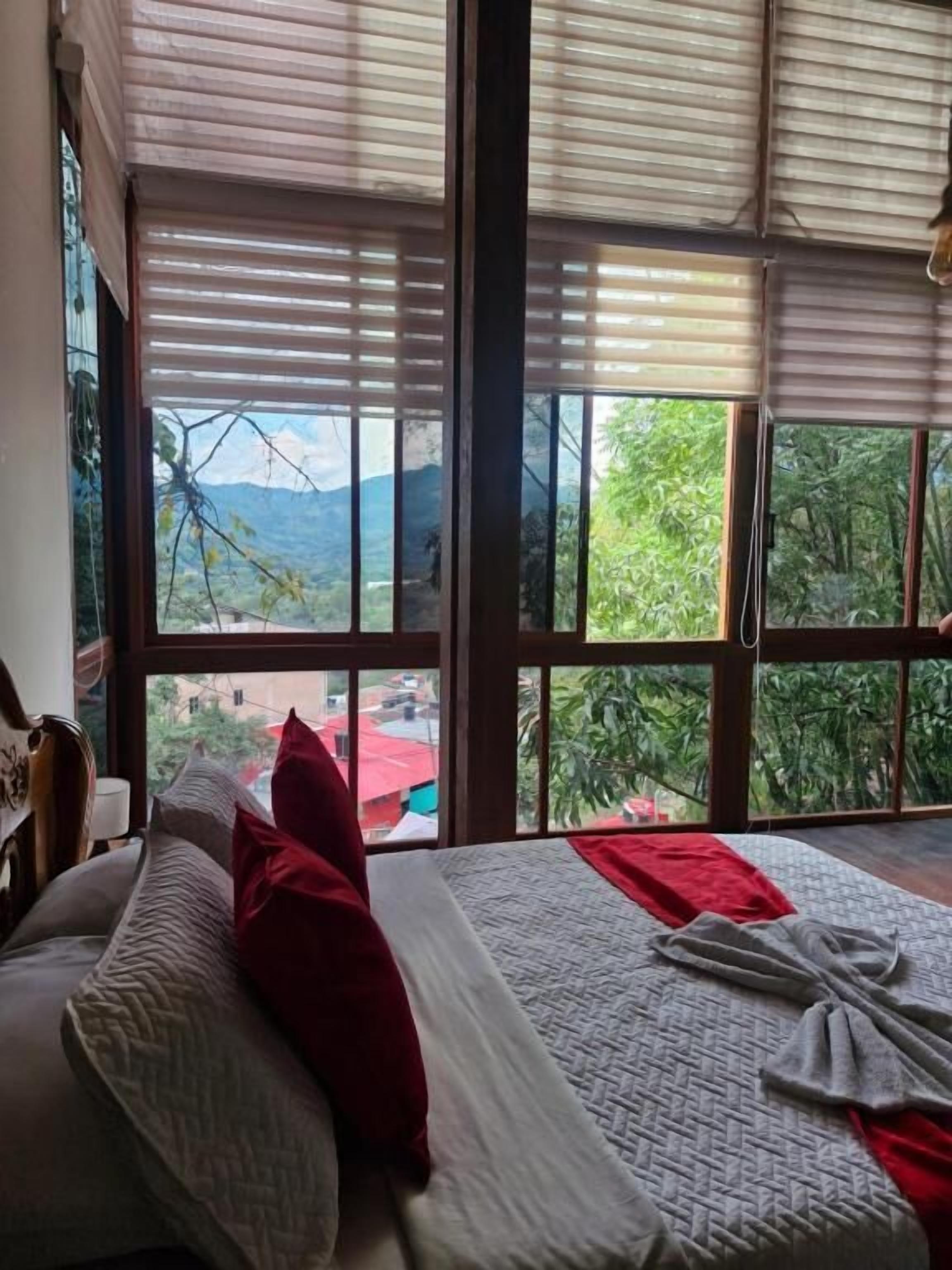 Quarto com cama, janelas amplas e vista para áreas verdes e montanhas.