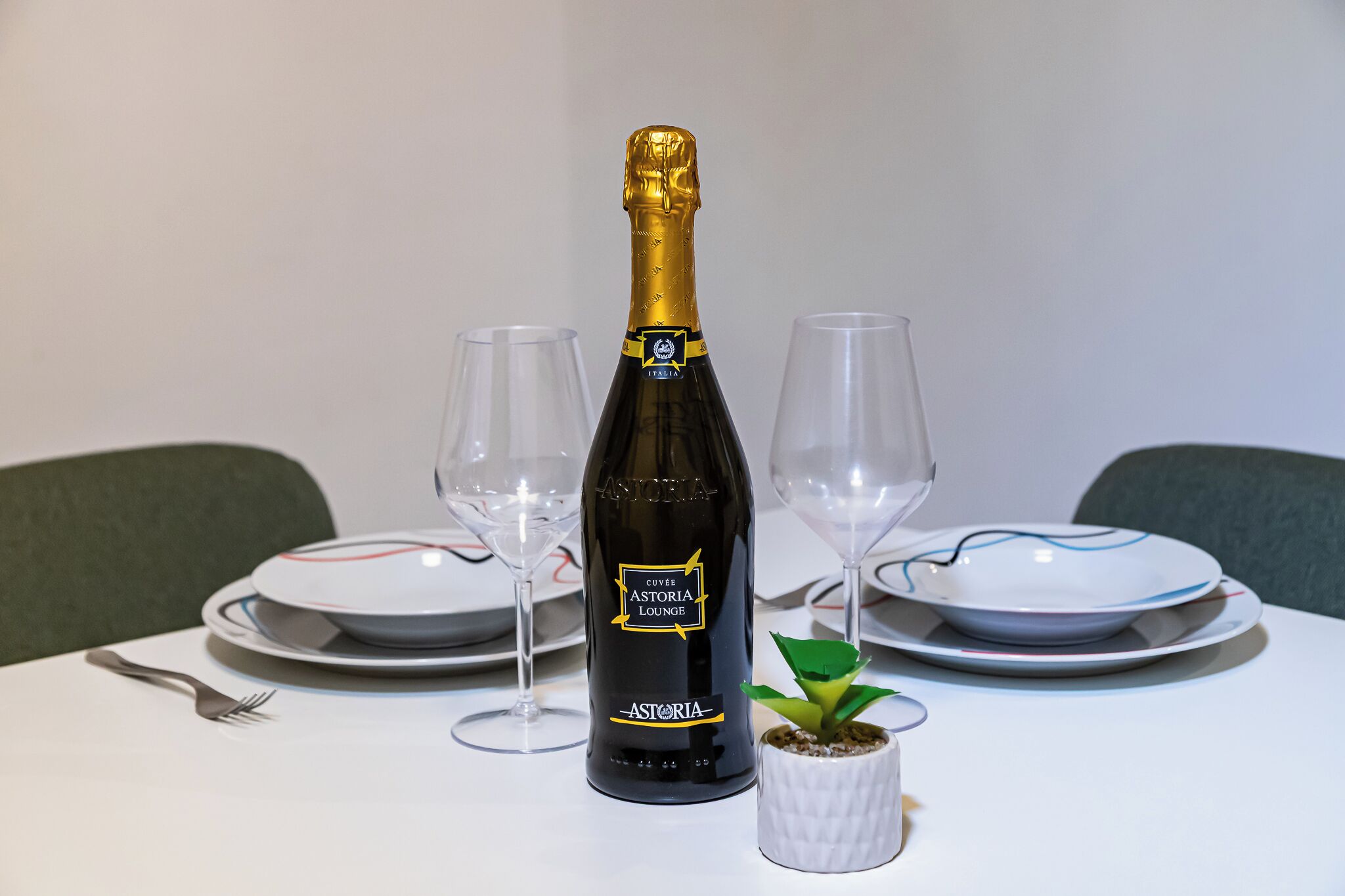 A bottle of Astoria champagne, two empty wine glasses, a small potted plant, and a fork on a table.