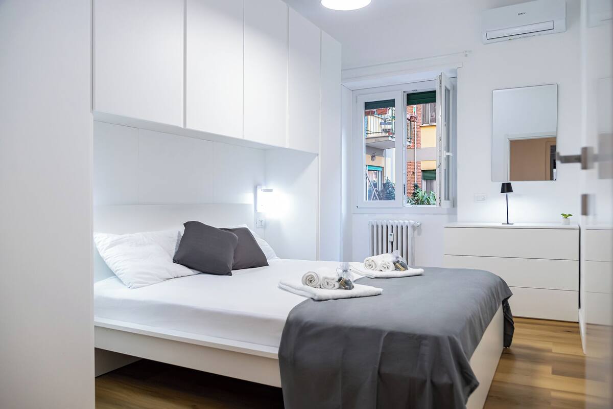 Une chambre d’hôtel moderne, équipée d’un lit, d’une commode, d’un miroir et d’une fenêtre donnant sur des immeubles.