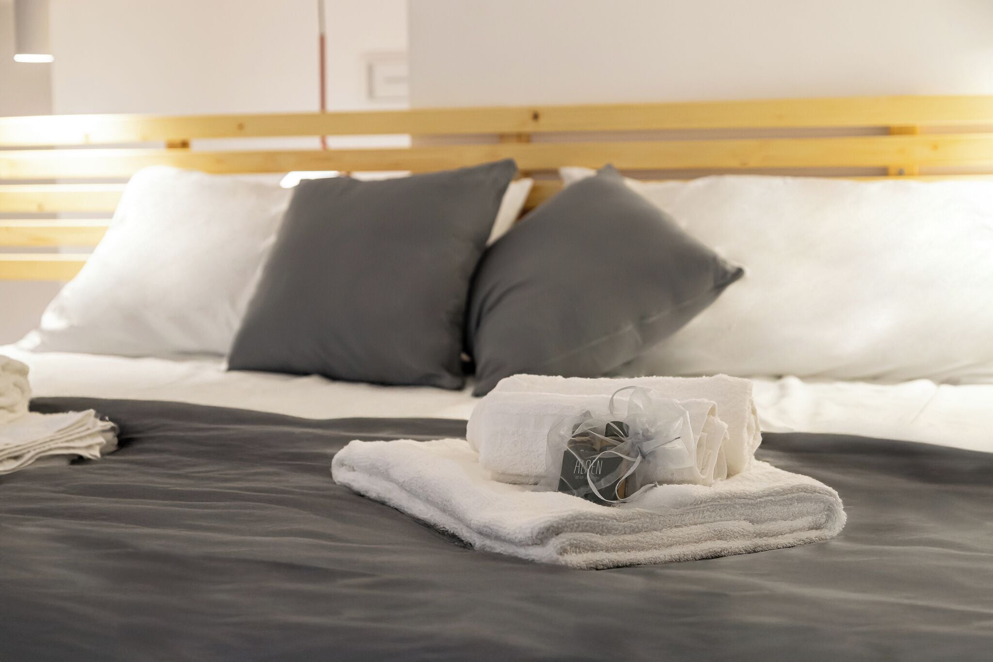 Ein ordentlich bezogenes Bett in Grau und Weiß, ein hölzernes Kopfbrett und ein gefaltetes Handtuch mit einem Etikett.