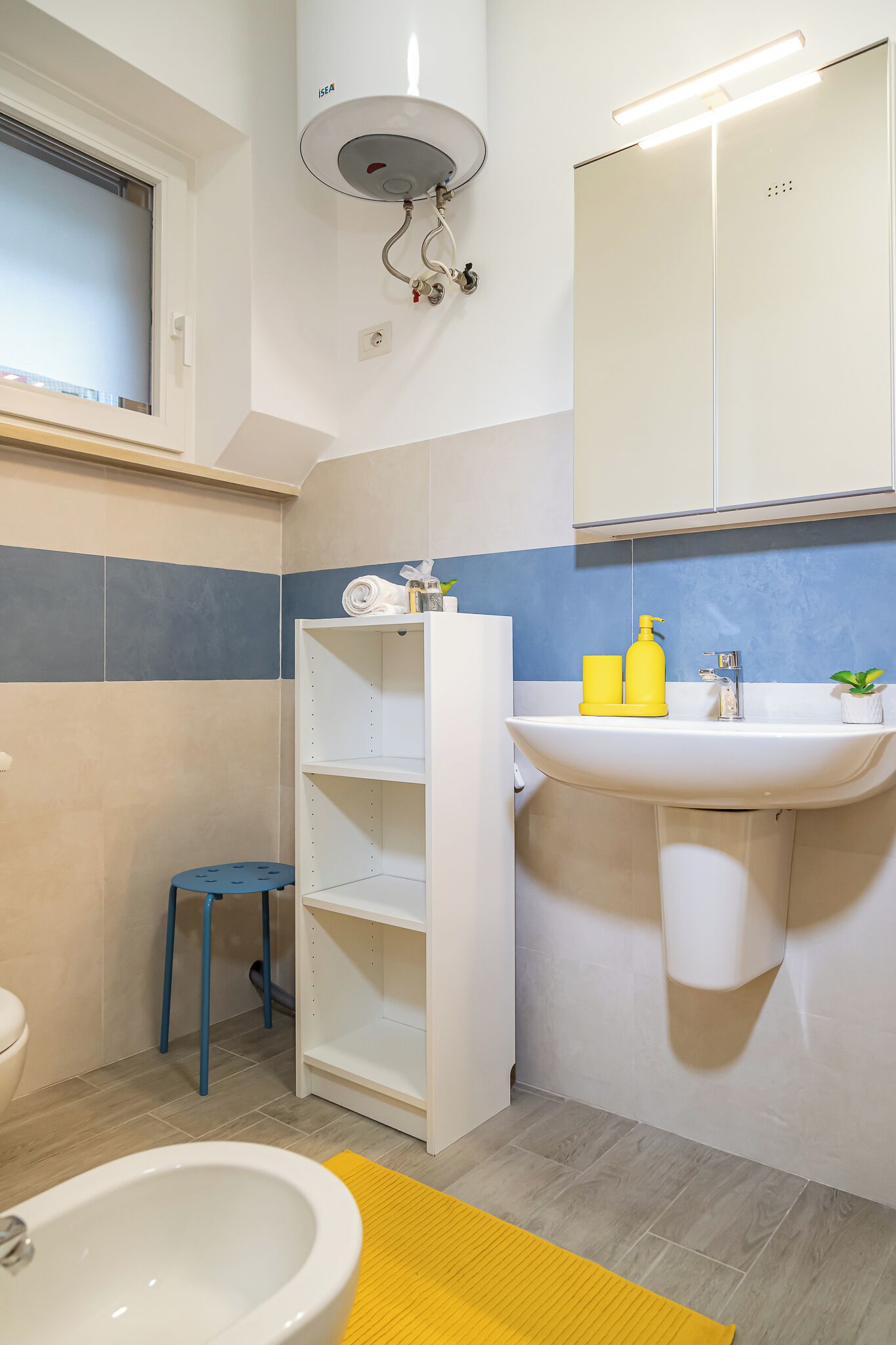 A bathroom with a toilet, bidet, white sink, and a shelf with toiletries.