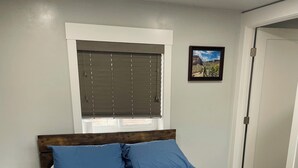A bedroom with a wooden bed, a quilted bedspread, a window with blinds, and a framed picture on the wall.