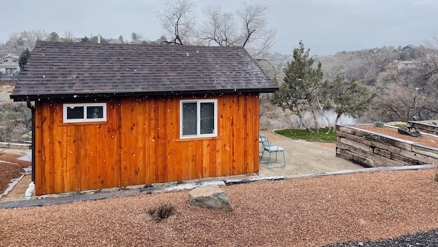 A small wooden cabin with a black roof, two windows, and a view of a lake and hills in the background.