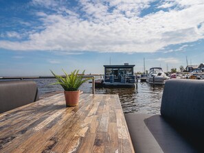 Una mesa de madera con una planta en maceta, un banco acolchado y vistas a un puerto deportivo con barcos y un pequeño edificio.