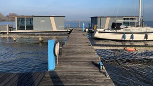 Une jetée en bois mène à une structure moderne flottante, dotée d’une façade en verre, et un voilier est amarré à proximité.