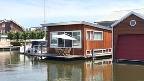 Ein modernes Hausboot mit einem großen Fenster, einer kleinen Terrasse mit Tisch und Stühlen und einem roten Garagengebäude.