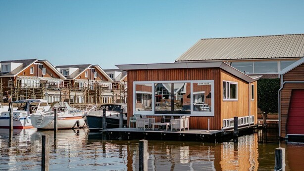 Ein modernes Holzhausboot liegt in einer Marina mit Booten vor Anker, der Himmel ist klar und die Wasseroberfläche ruhig.