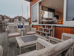 Terraza de madera con mobiliario exterior, vista a barcos y un edificio con un amplio ventanal e iluminación interior.