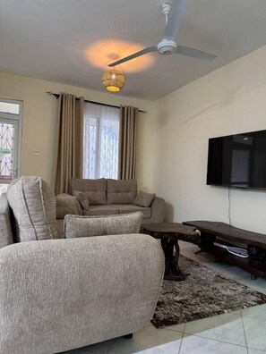 Ein Wohnzimmer mit einer Sofagarnitur, einem Deckenventilator, einem an der Wand montierten Fernseher und einem gemusterten Teppich.